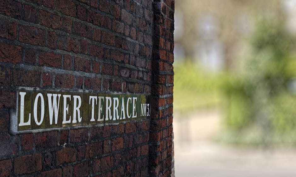 A close-up view of a brick wall featuring a street sign that reads 'LOWER TERRACE NW3', with the sign mounted onto the red bricks. The background is blurred, showing hints of greenery and trees illumi