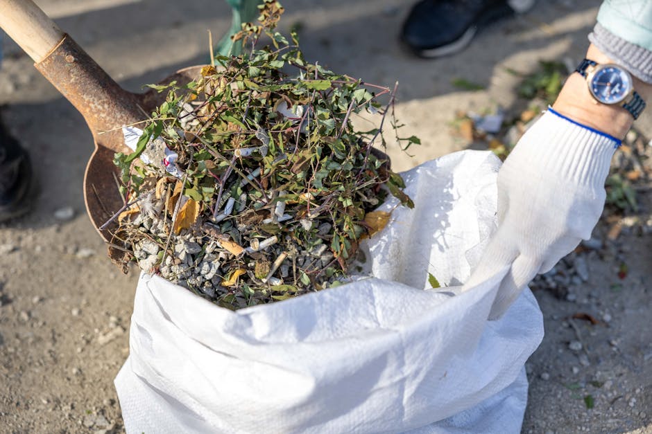 A person wearing a white gardening glove and a watch is using a shovel to lift a load of garden waste, including small branches, leaves, and gravel, into a large white sack. The sack is situated on an